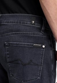 Dunkelblaue Jeans mit einem Etikett, auf dem die Zahl "7" zu sehen ist. Die Gesäßtasche hat ein Logo-Label und einen silbernen Knopf. Subtiler Steppnahtmuster.