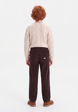 Enfant aux cheveux roux bouclés, de dos, portant un pull en maille torsadée crème, un pantalon marron foncé et des chaussures marron sur fond blanc.