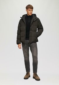 Dunkle gepolsterte Jacke mit Kapuze, ausgestattet mit einem Reißverschluss und seitlichen Taschen. Dazugehörig ein schwarzer Pullover mit Knopfleiste. Graue Jeans und braune Stiefel.