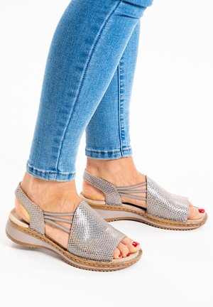 Voeten met beige sandalen met open tenen, elastische bandjes en een gestructureerd detail, gecombineerd met lichtblauwe opgerolde spijkerbroek, op een witte achtergrond.