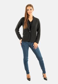 Zwarte blazer met een sjawlkrans, enkele knoopsluiting en twee voorzakken, gecombineerd met strakke jeans en zwarte hoge hakken.