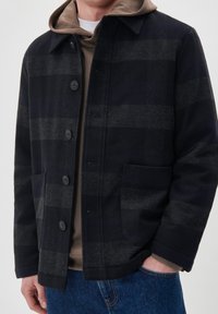 Chaqueta de mezcla de lana de color azul marino con patrón de cuadros grises, que presenta un cierre de botones en la parte delantera, dos bolsillos grandes y un collar puntiagudo. Lleva puesta una sudadera marrón debajo.