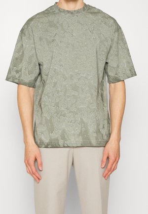 T-shirt oversize vert olive avec un motif texturé et irrégulier, col rond et manches courtes, présenté sur un fond blanc.