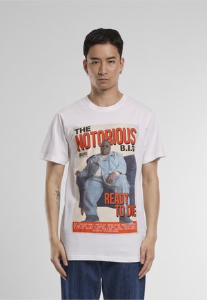 Homme portant un T-shirt blanc avec l'impression de la pochette de l'album "Ready to Die" de The Notorious B.I.G., jean bleu, debout devant un fond gris uni.