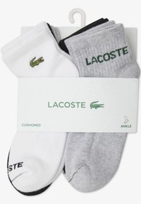 Ankle socks in white and gray, featuring a green Lacoste logo and cushioned texture. Packaged with a hanging tag.