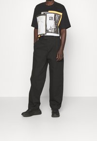 Svart grafisk t-shirt med blandade färger på ramar och bilder, kombinerad med lösa svarta byxor och svarta sneakers.