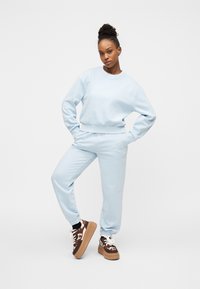 Νεαρή γυναίκα στέκεται με τα χέρια στις τσέπες, φορώντας ανοιχτό μπλε φούτερ και φόρμες, με καφέ και λευκά παπούτσια πλατφόρμας.