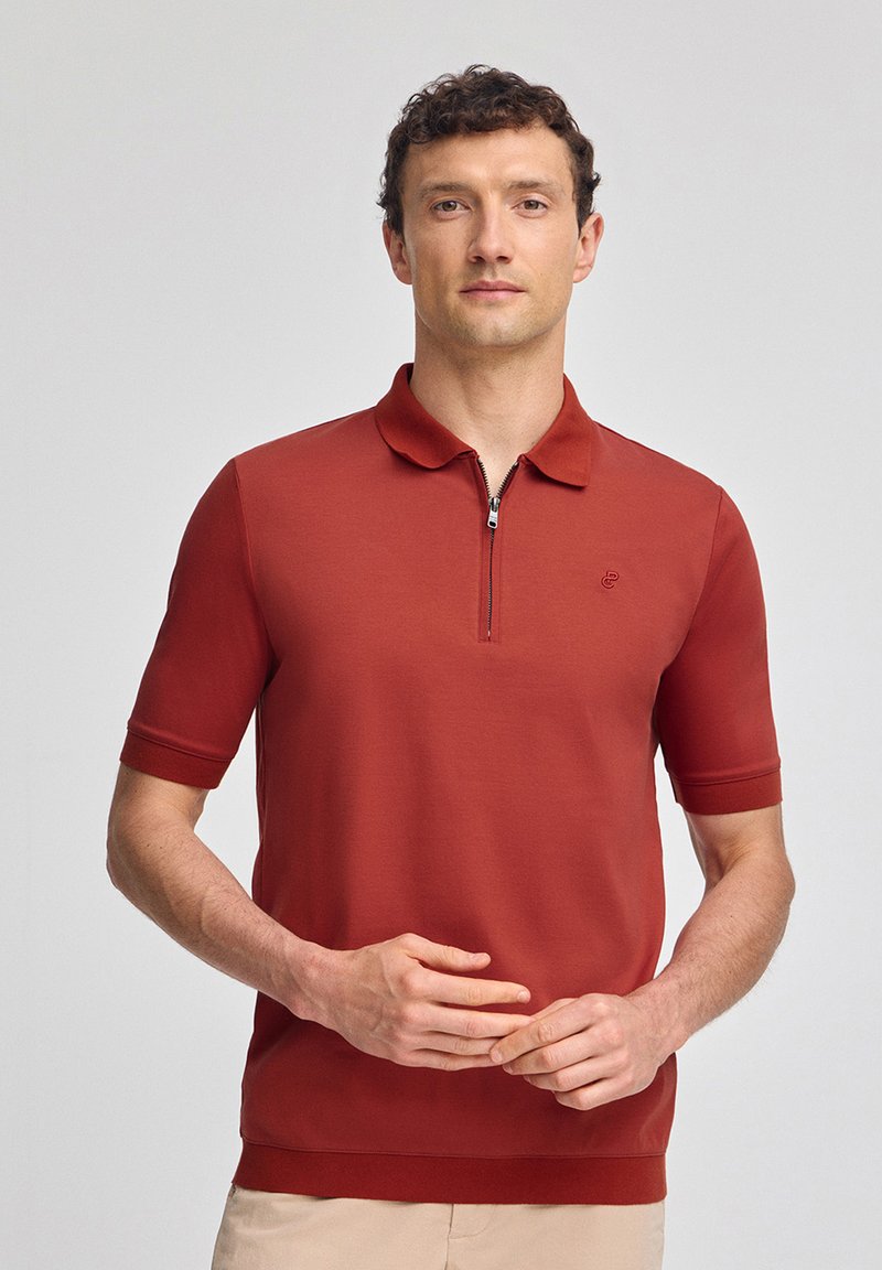 Kortärmad pikétröja i rostbrun med dragkedja i halsen, ribbad krage och muddar, med en liten logotyp på bröstet.