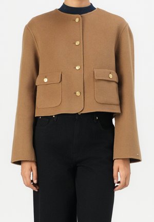 Brązowa krótka kurtka na guziki z dwoma przednimi kieszeniami z klapkami, noszona na czarnym topie i czarnych spodniach z wysokim stanem.