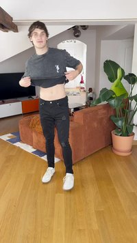 Čierne roztrhnuté úzke džínsy, šedé skrátené tričko s logom, stojí na drevenej podlahe v obývacej izbe s izbovými rastlinami a pohovkou.