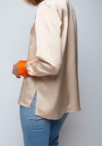 Silk blouse in beige with long sleeves; features orange cuffs and side slits. Paired with light blue denim for contrast.