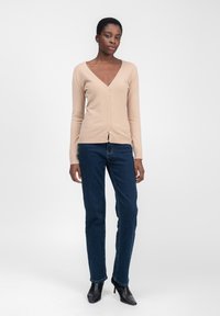 Beige Strickjacke mit V-Ausschnitt, Knopfverschluss und langen Ärmeln, kombiniert mit dunkelblauen Jeans und schwarzen spitzen High Heels.