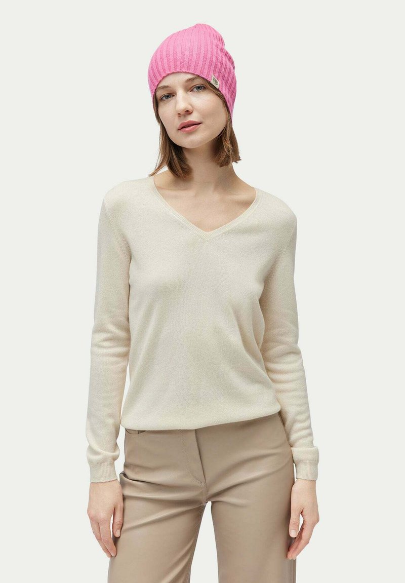 Krämfärgad v-ringad tröja i mjukt stickat tyg, parat med en ribbad rosa beanie. Kvinnan har axellångt hår och ljusbeiga byxor.