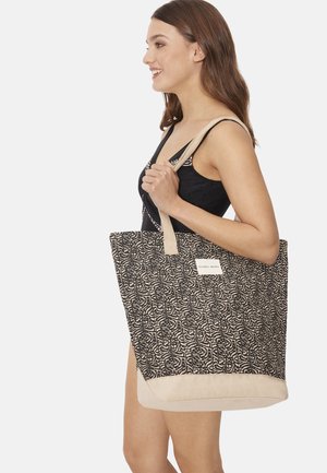 Mujer con traje de baño negro sostiene un bolso grande de mano con estampado beige y negro y asas beige sobre su hombro, sonriendo hacia la derecha.