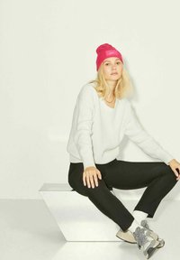 Suéter branco canelado, calças pretas e um gorro rosa brilhante. Ténis cinzentos com detalhes. Sentado em um pedestal geométrico. Fundo simplista.