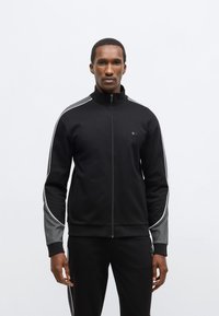 Čierna bunda na zips s vysokým golierom, sivé akcenty na pleciach a biele lemovanie. Vyrobená z mäkkého materiálu, priliehavý strih s vreckami.