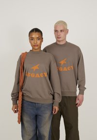 Sweatshirt maron cu text portocaliu „LEGACY” și grafică cu stea. Material din bumbac și alte fibre, croială relaxată, manșete și tiv ribbate. Modelul ține o geantă țesută.