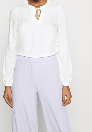 Camicia bianca con scollatura rotonda, dettaglio con fiocco davanti e maniche lunghe a palloncino. Abbinata a pantaloni larghi plissettati di color lavanda chiaro.