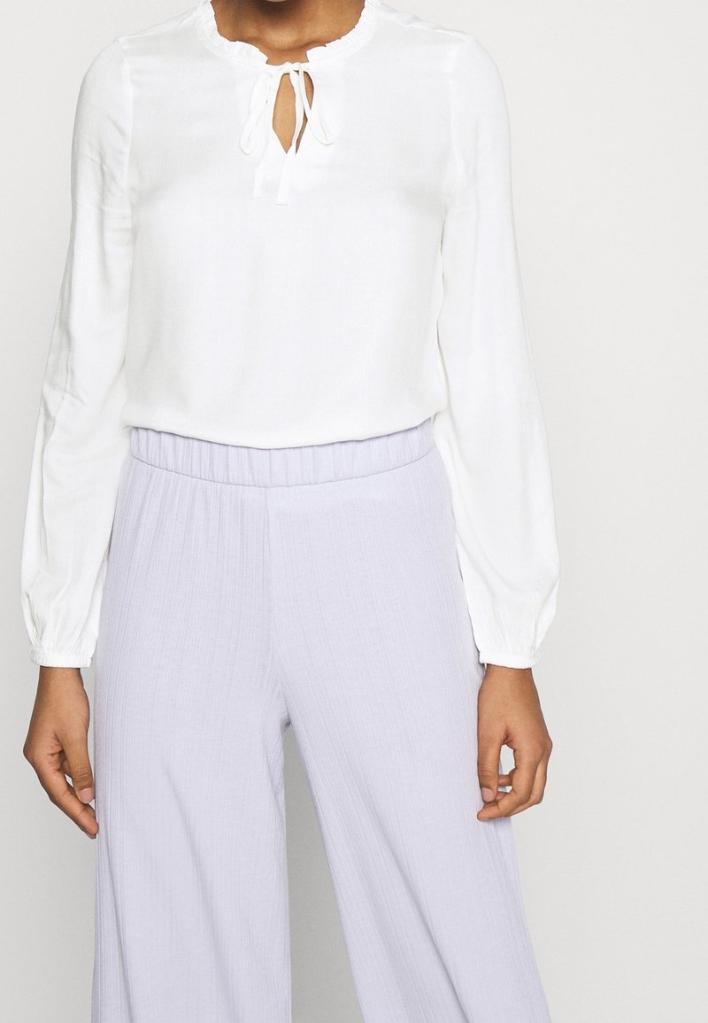 Camicia bianca con scollatura rotonda, dettaglio con fiocco davanti e maniche lunghe a palloncino. Abbinata a pantaloni larghi plissettati di color lavanda chiaro.