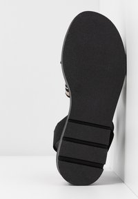 Semelle de sandale noire avec trois rainures horizontales, présentée à la verticale contre un fond de mur blanc en angle.