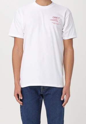 Camiseta de algodón blanca con cuello redondo, mangas cortas y un estampado gráfico rojo que presenta la palabra "OBEY" y un diseño en espiral en la parte frontal.