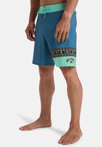 Bañadores de hombre en azul y verde menta, con una banda estampada y logotipo en negro. Fabricados con material ligero y de secado rápido.