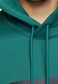 Tummansininen huppari, jossa on teksturoitu kangas, varustettuna kiristysnyörillä ja tummemmalla sävyllä oleva graafinen yksityiskohta rinnassa.