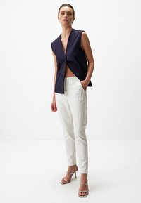 Marineblauw mouwloos topje met een diepe V-hals en een plooidetail aan de voorkant, gecombineerd met witte gestreepte tailored broek en zilveren sleehak sandalen. Minimalistisch ontwerp.