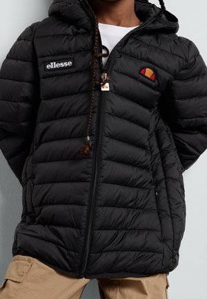 Bambino con giacca trapuntata nera con cappuccio e patch logo ellesse sul petto e pantaloni cargo beige, in piedi contro una parete semplice.