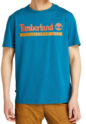 Blå kortærmet skjorte med orange "Timberland Established 1973" tekst og trælogo, båret med bukser i sennepsfarve.