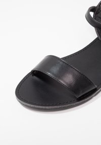 Musta nahkasaapas, jossa on kaksi leveää hihnaa, sileä pinta, avoin kärki ja litteä pohja, joka korostaa minimalistista rakennetta ja ompelutietoja.