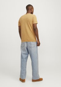 Camiseta de manga corta, de color mostaza, con una textura suave, combinada con unos vaqueros sueltos de color azul claro y calzado marrón, vista desde la espalda.