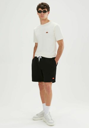 Junger Mann trägt weißes T-Shirt, schwarze Shorts mit Kordelzug, weiße Sneakers, Socken und schwarze Sonnenbrille, steht mit Händen in den Taschen.