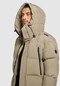 Chaqueta puffy beige con una gran capucha, cremalleras negras y un logo redondo. Presenta un material texturizado con costuras horizontales para mayor calidez.
