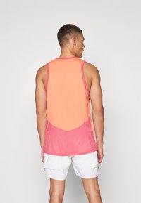Canottiera in rete con una sfumatura da arancione a rosa, dal taglio sportivo e dettagli di cucitura a vista. Abbinata a pantaloni corti bianchi.