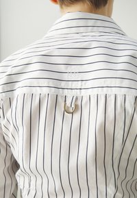 Rutet skjorte i hvit og marine, med klassisk krage, detalj med løkke bak, og dekorativt brodert merke ved halsen. Glatt tekstur.