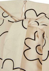 Tissu crème avec un motif de fleurs à contour noir, présente un détail de nœud à la taille et une fermeture éclair.