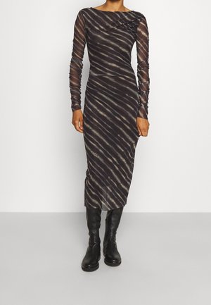 Vrouw die een midi-jurk met lange mouwen draagt, zwart en grijs gestreept, met doorschijnende mouwen en zwarte kniehoge laarzen, staand voor een effen achtergrond.