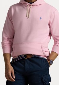 Rosa hoodie med en framficka och dragsko på huvan. Har en liten blå logotyp på bröstet. Kombinerad med mörka byxor och en klocka.