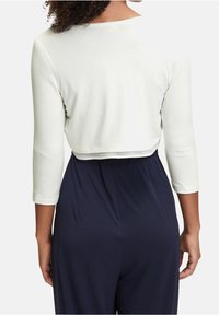 Mujer con una blusa blanca de manga larga corta y pantalones de tiro alto de color azul oscuro, vista desde atrás frente a un fondo sencillo.