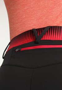 Un pantalon de sport noir présente une taille haute avec une ceinture à motifs rouges et noirs et une petite poche zippée à l'arrière.