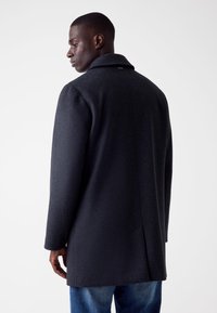 Cappotto grigio scuro con colletto strutturato, maniche lunghe e linee pulite. Il tessuto appare testurizzato, completato da un design semplice sul retro.