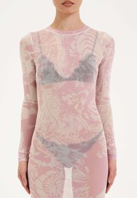 Průhledný top s dlouhým rukávem s růžovým a krémovým květinovým vzorem. Má kulatý výstřih a přiléhavý střih, přičemž je vrstvený přes šedou podprsenku.