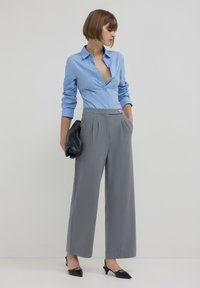 Hellblaues Hemd mit langen Ärmeln, kombiniert mit grauen, weit geschnittenen Hosen, die ein Knopfdetail aufweisen. Schwarze spitze Schuhe und eine Clutch.