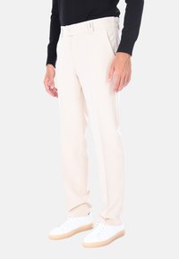 Uomo che indossa pantaloni beige su misura e sneaker bianche, in piedi contro uno sfondo chiaro e uniforme con le mani rilassate.