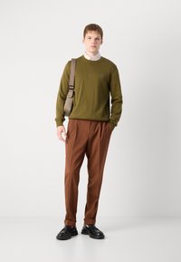 Olivgrüner Pullover mit gestrickter Textur, braune plissierte Hose und schwarze Schuhe. Der Taschengurt überquert den Körper; kleines Logo auf der Brust des Pullovers.