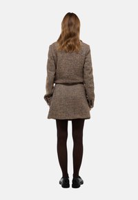 Ensemble de manteau et jupe en laine mélangée, avec un motif en chevrons marron, doté de longues manches, d'une taille cintrée, associé à des collants foncés et des chaussures noires.