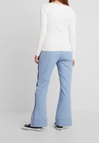 Personne portant un pantalon évasé à boutons latéraux bleu clair, un haut blanc à manches longues côtelé et des baskets Converse noires, debout sur un fond blanc.