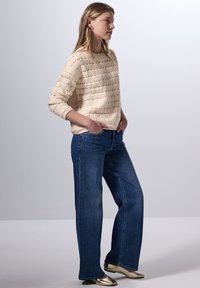 Knitcreme Pullover mit einem strukturierten Stichmuster, kombiniert mit weit geschnittenen blauen Jeans und metallic-goldenen spitzen Schuhen.
