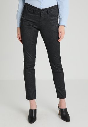 Mujer con pantalones negros ajustados, camisa azul claro abotonada y mules negras de tacón, de pie frente a un fondo liso.